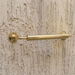 May include: A brass towel rail fixed to a textured wall. The rail is cylindrical with circular mounting plates at each end. The polished brass finish gives a vintage appearance. The rail's length is approximately 30 cm.
