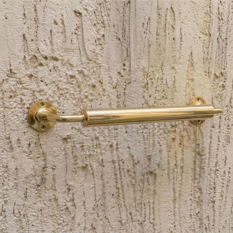 May include: A brass towel rail fixed to a textured wall. The rail is cylindrical with circular mounting plates at each end. The polished brass finish gives a vintage appearance. The rail's length is approximately 30 cm.