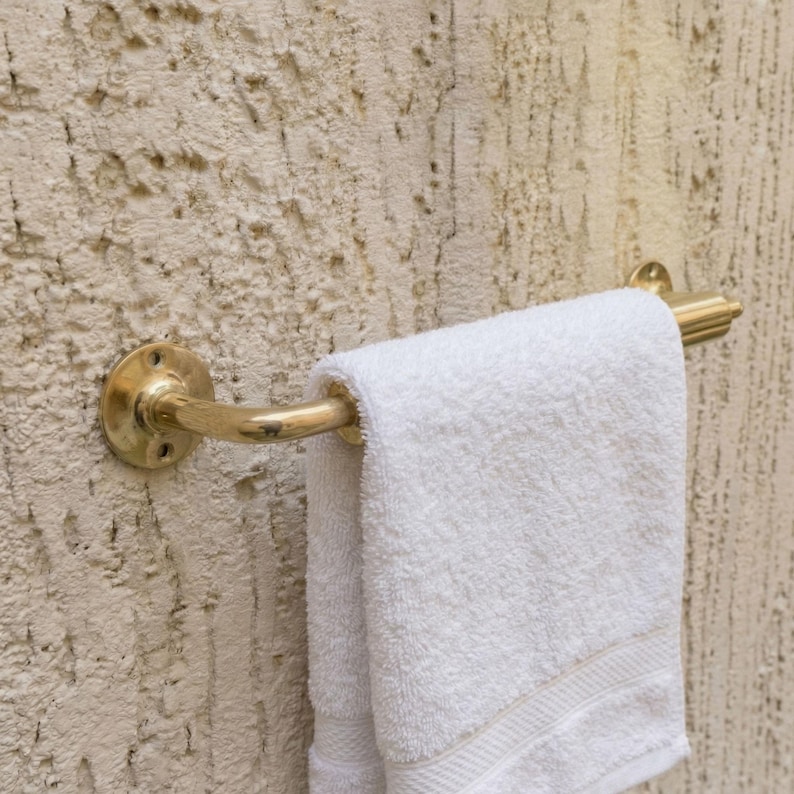 May include: A brass towel rail fixed to a textured wall, holding a folded white towel. The rail has a curved design with decorative detailing. The wall has a rough, stucco-like finish.