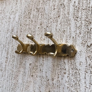 Pode incluir: Um cabide de parede de cor latão com três ganchos. Os ganchos têm um topo arredondado e um design curvo. O cabide está montado numa parede texturizada esbranquiçada. O cabide tem cerca de 25 cm de comprimento.