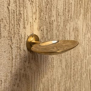 May include: A brass-colored door handle with a circular base and a curved, oval-shaped handle. The handle is mounted on a textured, light brown wall. The handle's design is simple and elegant, suitable for various interior styles.