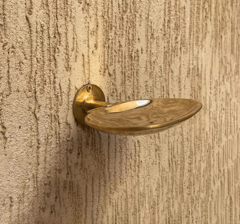 May include: A brass-colored door handle with a circular base and a curved, oval-shaped handle. The handle is mounted on a textured, light brown wall. The handle's design is simple and elegant, suitable for various interior styles.