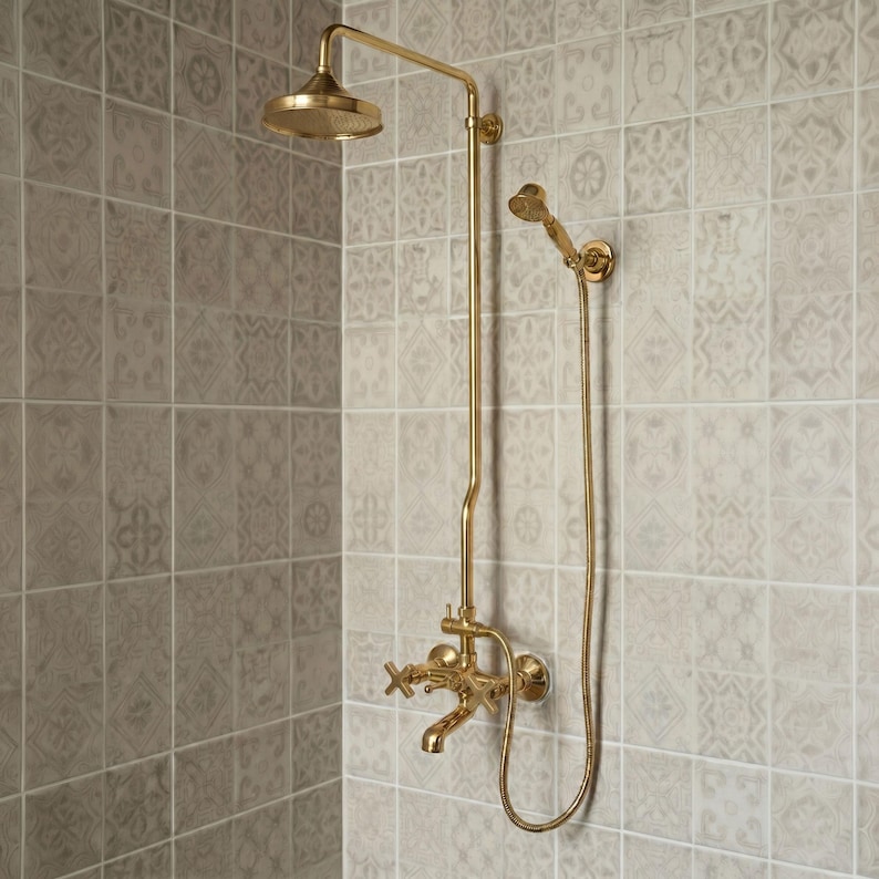 May include: A gold-toned shower system featuring a fixed overhead shower head, a flexible handheld shower head with a hose, and a wall-mounted faucet with two cross-style handles. The fixture is mounted against a wall of light gray patterned tiles.