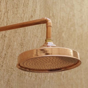 May include: A copper shower head with a round, perforated spray plate. The shower head is attached to a copper pipe and elbow joint. The fixture has a polished finish and is set against a textured, light brown wall.