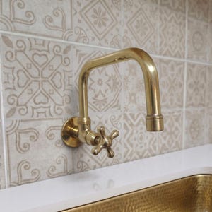 Könnte beinhalten: Ein wandmontierter Messingwasserhahn mit hohem Auslauf und einem vierarmigen Kreuzgriff. Die Armatur hat eine polierte metallische Oberfläche und ist über einer weißen Arbeitsplatte und einem gehämmerten Messingwaschbecken angebracht.