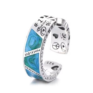 Puede incluir: Anillo ajustable de color plateado con una incrustación de esmalte azul y verde veteado. El anillo está grabado con las palabras "PEACE LOVE" y otros símbolos. El anillo está marcado con "925" y otro texto.