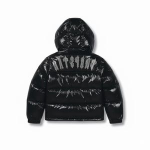 Può includere: Giacca trapuntata nera con cappuccio. La parte posteriore della giacca presenta la scritta "TRAPSTAR" in argento, con un carattere in stile gotico. La giacca ha una finitura lucida.