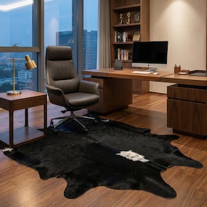 May include: A contemporary home office with a dark brown desk, matching shelving, and a black leather chair. A black and white cowhide rug lies on the hardwood floor. A gold desk lamp and a side table are also present.
