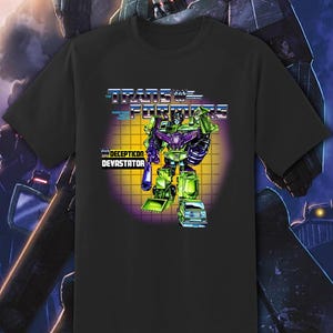 May include: Black t-shirt featuring a graphic of the Transformers character Devastator. The design includes the Transformers logo and the text "DECEPTICON DEVATASTOR". The robot is depicted in green and purple with a yellow and orange background.