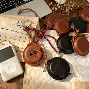 以下が含まれることがあります： 革製のアクセサリーのコレクション。丸いケースにはコードが取り付けられ、栓抜きも含まれています。ケースは茶色と黒の色合いで、一部にはエンボス加工のデザインと「Anabel」という名前が施されています。iPodも写っています。
