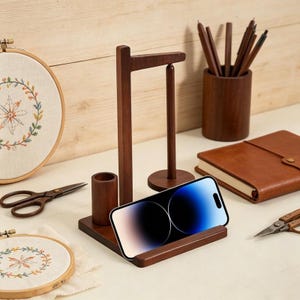以下が含まれることがあります： 濃い茶色の木製電話スタンド。ペンホルダーとスプールホルダー付き。スマートフォンがスタンドに置かれています。革製ジャーナル、刺繍枠、ハサミ、ペンなどのアイテムも含まれています。