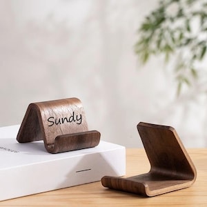 以下が含まれることがあります： 2つの木製電話スタンド。1つのスタンドは角度がついており、「Sundy」の文字が印刷されています。スタンドは濃い茶色の木材でできており、電話やタブレットを保持するように設計されています。スタンドは明るい色の表面にあります。