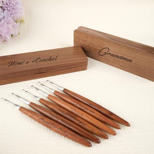 以下が含まれることがあります： 銀色の金属製の先端が付いた6本の木製かぎ針編みフックのセット。それぞれに名前またはメッセージが刻まれています。フックは、「Mimi's Crochet」と「Grandma」と書かれた2つの木箱と一緒に表示されます。