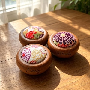 以下が含まれることがあります： 木製のピンクッション3つ。それぞれ赤、紫、黄色の花柄の布地が使われています。「Emily」と「Mandy」の名前が2つのクッションに刻まれています。