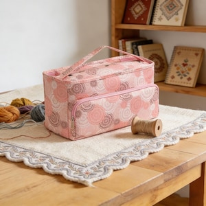 以下が含まれることがあります： ピンクの花柄の糸収納バッグ。ハンドルとフロントジッパーポケット付き。バッグは、糸と木製のスプールが近くにある、クリーム色の刺繍マットの上に置かれています。編み物やかぎ針編みの用品を収納するように設計されています。