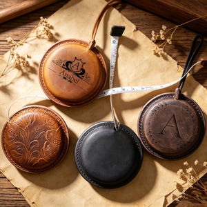 以下が含まれることがあります： 茶色と黒の色合いの丸い革製メジャーテープ4つ。白いメジャーテープが伸びています。1つは「Anabel」という名前とロゴ、もう1つは花の模様、3つ目は文字「A」が描かれています。