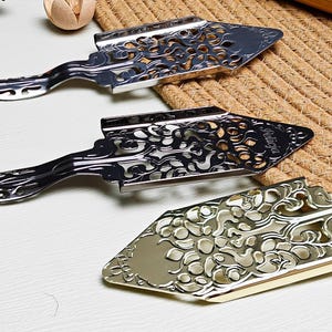 May include: Close-up of three decorative metal pie servers. The servers have ornate, openwork designs and long handles. One is silver, one is black, and one is gold. The servers are on a white surface.