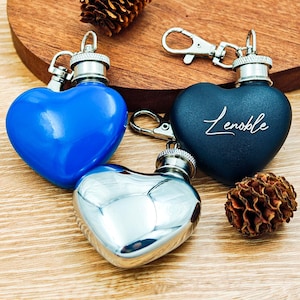 Op de afbeelding: Hartvormige heupflacons in blauw, zilver en marineblauw met zilveren hardware. Eén marineblauwe flacon heeft de naam "Lenoble" in cursief. De flacons liggen op een houten oppervlak met een dennenappel.