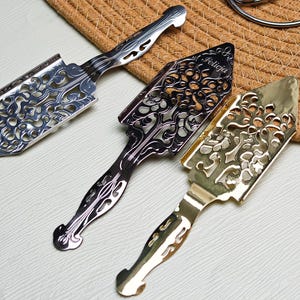 May include: Three decorative absinthe spoons with ornate, openwork designs. One is silver, one is black, and one is gold. The spoon in the middle has the name "Felicia" engraved on it. The spoons are arranged on a light surface.