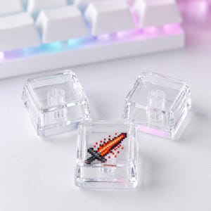 May include: Three square, transparent mechanical keyboard keycaps. Two keycaps are clear, while the center keycap features a small, detailed pixel art graphic of a flaming sword with orange, red, and black colors on its top surface.