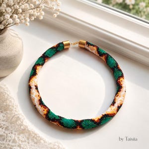 May include: A beaded necklace with a green, white, black, and brown pattern. The necklace has gold-colored clasps and is displayed on a white surface. The necklace is a handmade jewelry item.