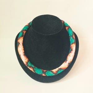 May include: A beaded necklace with a vibrant pattern of green, orange, and peach colors. The necklace is displayed on a black velvet bust, showcasing its circular design and intricate beadwork. The beads are small and tightly strung.