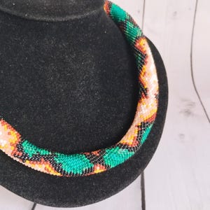 Puede incluir: Un collar de cuentas de colores, exhibido sobre un busto negro. El collar presenta un diseño tubular con un patrón repetido de cuentas verdes, naranjas, negras y blancas. Las cuentas están bien ensartadas, creando una superficie lisa y texturizada.