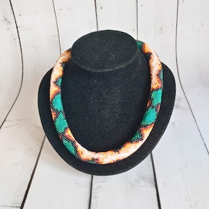 Könnte beinhalten: Eine Perlenkette, präsentiert auf einer schwarzen Büste. Die Halskette hat ein Muster aus grünen, orangefarbenen und schwarzen Perlen auf cremefarbenem Grund. Die Halskette ist röhrenförmig und scheint handgefertigt zu sein.