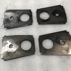 May include: Four dark metal plates with a rectangular shape, each featuring a large circular hole and a smaller hexagonal hole. The plates have a worn, industrial appearance with visible scratches and discoloration.
