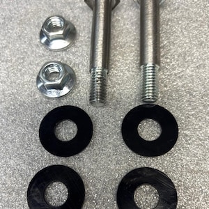 May include: An assortment of hardware components, including two silver bolts with hexagonal heads, two silver nuts, and four black rubber washers. The components are arranged on a textured gray surface.