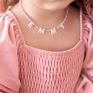May include: A pink beaded necklace spelling out the name "EMMY" in white block letters. The necklace is worn by a person wearing a pink dress with a square neckline and puffed sleeves. The dress has a textured bodice.