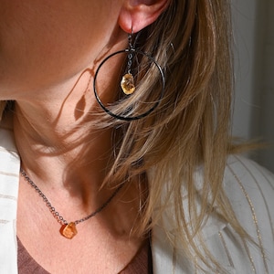 May include: A woman wearing a white blazer with gold stripes and a brown top. She is wearing a delicate necklace with a small, orange gemstone pendant. She is also wearing a pair of black hoop earrings with a dangling orange gemstone.