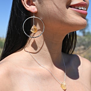 May include: A silver necklace and earrings set featuring a raw, yellow citrine stone pendant and matching earrings. The necklace is a delicate silver chain with a simple design. The earrings are hoop-style with a dangling citrine stone.