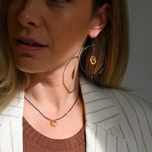 May include: A woman wearing a white striped blazer with a brown top and a delicate gold necklace with a small orange stone pendant. She is also wearing black hoop earrings with a small orange stone accent.