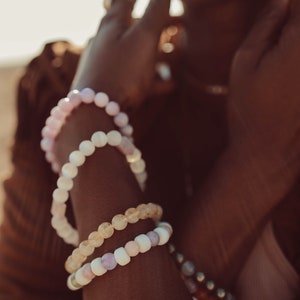 May include: A person wearing a stack of beaded bracelets on their wrist. The bracelets are made of different colored stones, including pink, white, and yellow.