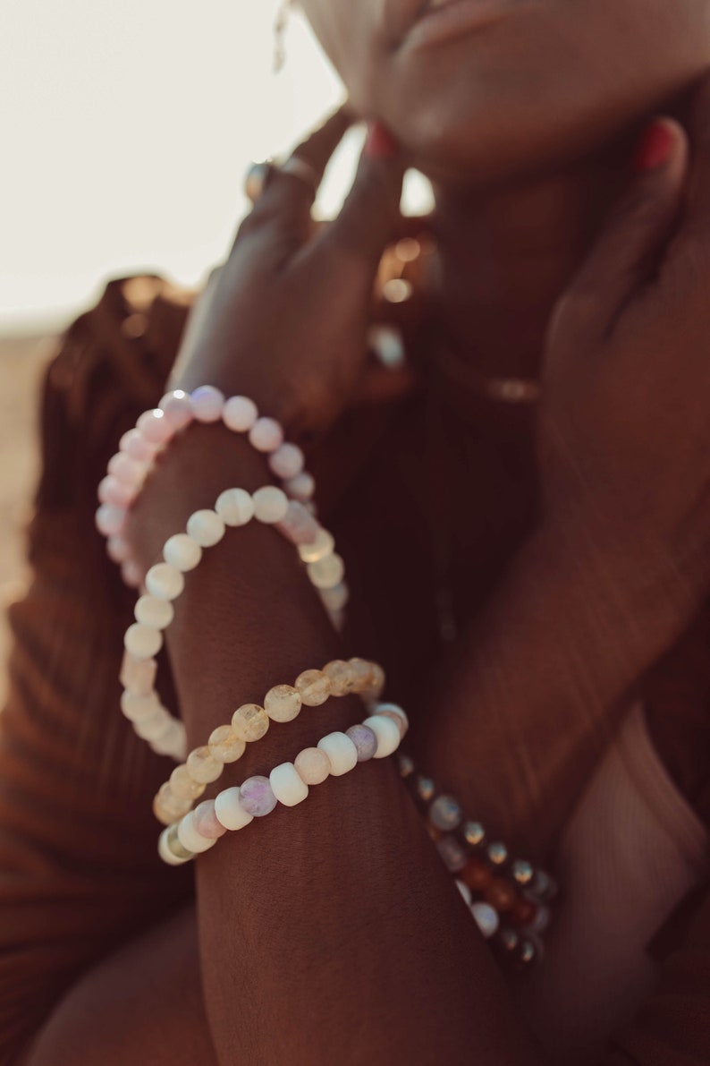 May include: A person wearing a stack of beaded bracelets on their wrist. The bracelets are made of different colored stones, including pink, white, and yellow.