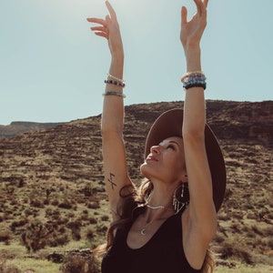 May include: A woman wearing a black tank top, a wide-brimmed hat, and multiple bracelets on both wrists, stands with her arms raised in the air. She is looking up at the sun, which is shining brightly in the sky. The background is a desert landscape with hills and shrubs.