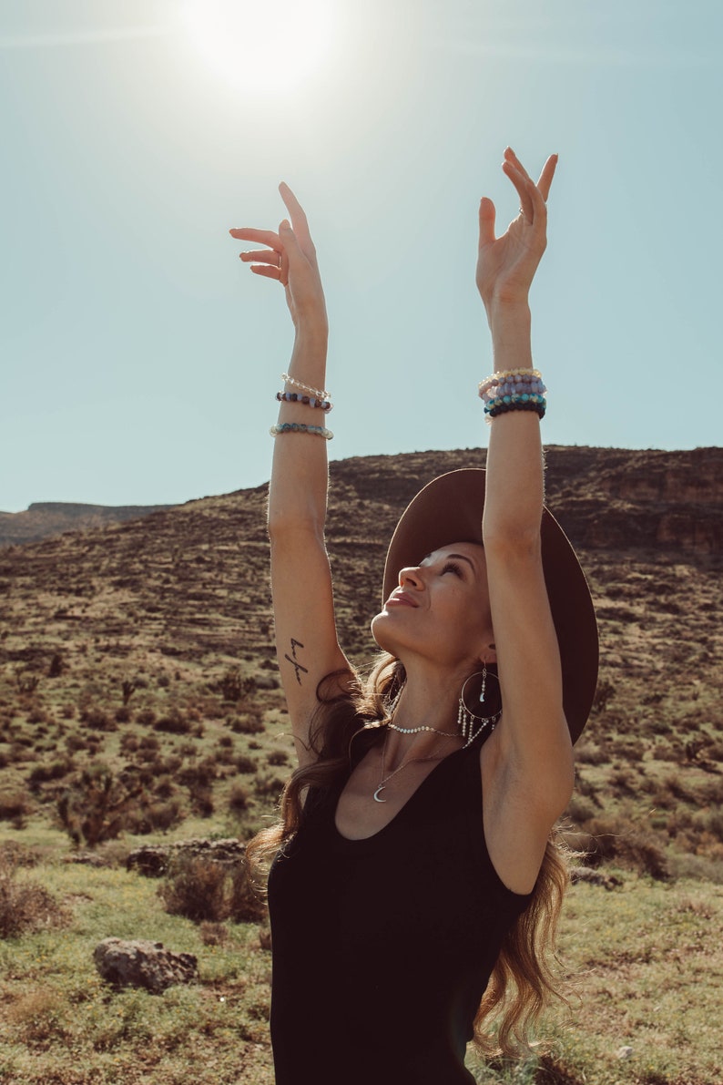 May include: A woman wearing a black tank top, a wide-brimmed hat, and multiple bracelets on both wrists, stands with her arms raised in the air. She is looking up at the sun, which is shining brightly in the sky. The background is a desert landscape with hills and shrubs.