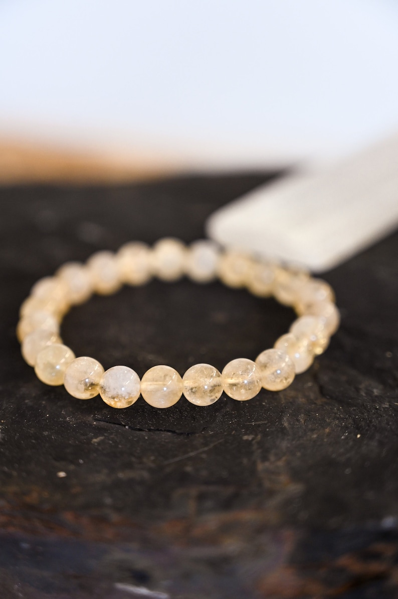 May include: A bracelet made of light yellow, translucent, round beads. The bracelet is on a dark brown surface.