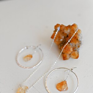 May include: A pair of silver hoop earrings with a dangling, raw, orange-colored gemstone. The earrings are on a white surface with a cluster of orange crystals in the background.