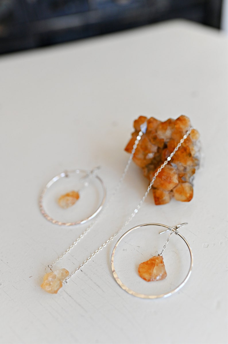 May include: A pair of silver hoop earrings with a dangling, raw, orange-colored gemstone. The earrings are on a white surface with a cluster of orange crystals in the background.