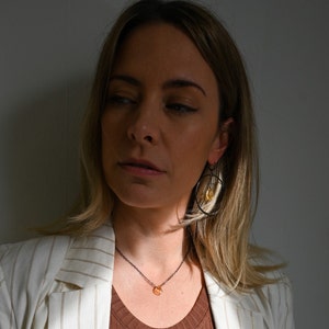 May include: A woman wearing a white striped blazer with a brown tank top underneath. She is wearing a delicate necklace with a small orange stone pendant and black hoop earrings with a small orange stone hanging from one.