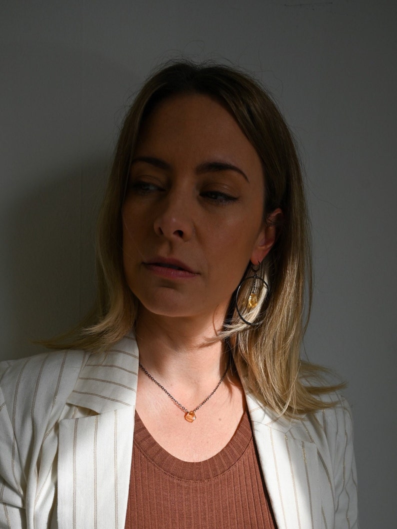 May include: A woman wearing a white striped blazer with a brown tank top underneath. She is wearing a delicate necklace with a small orange stone pendant and black hoop earrings with a small orange stone hanging from one.