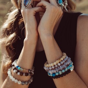 May include: A woman wearing a brown hat and a black top with multiple bracelets made of different colored stones. The bracelets are stacked on her wrist.