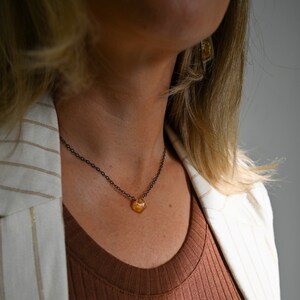 May include: A woman wearing a brown top and a white blazer with black stripes. She is wearing a delicate necklace with a small, orange, faceted gemstone pendant.