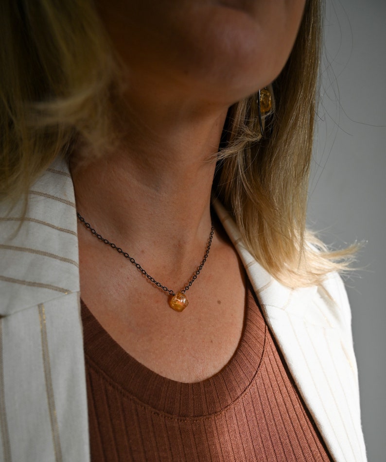 May include: A woman wearing a brown top and a white blazer with black stripes. She is wearing a delicate necklace with a small, orange, faceted gemstone pendant.
