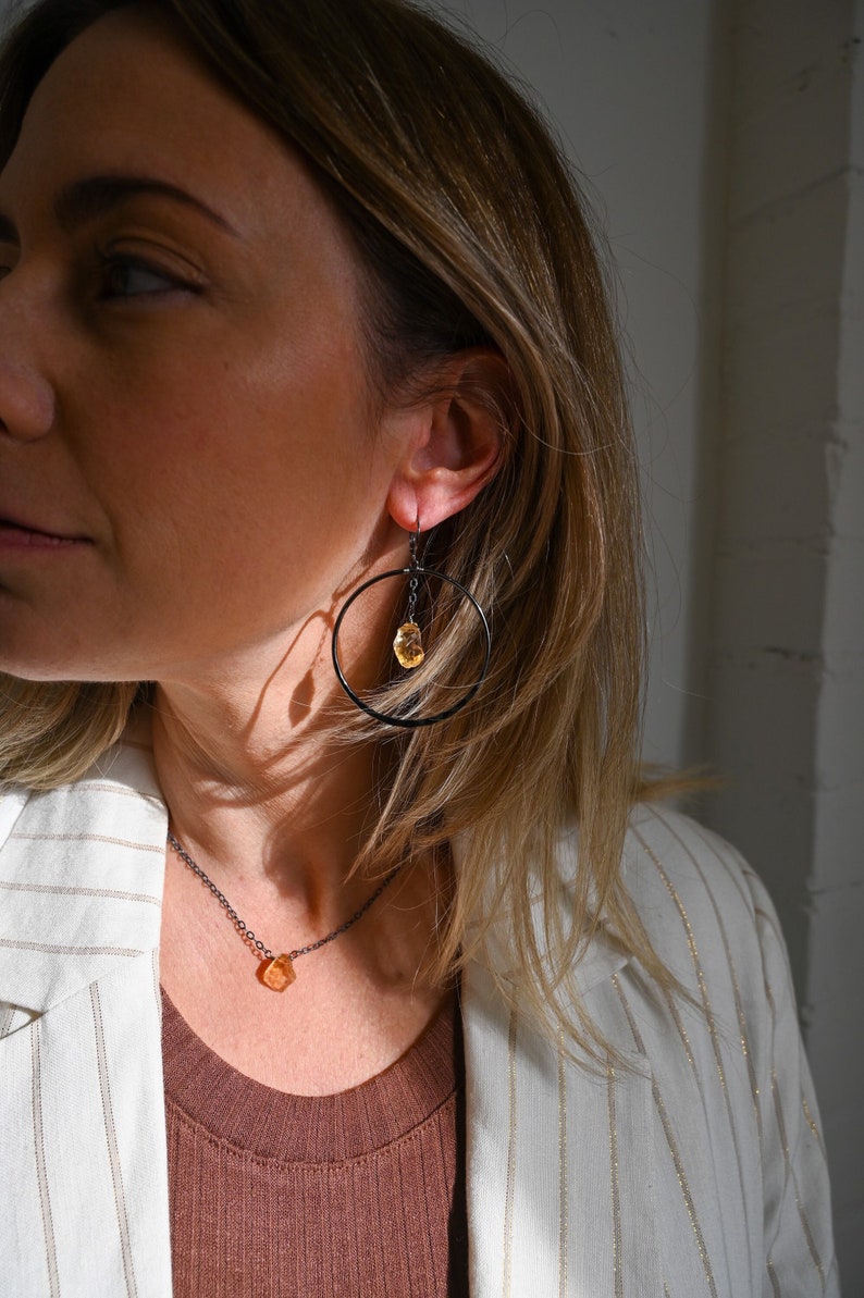 May include: A woman wearing a white blazer with gold stripes and a brown top. She is wearing a delicate necklace with a small, orange gemstone pendant. She is also wearing a pair of black hoop earrings with a dangling orange gemstone.