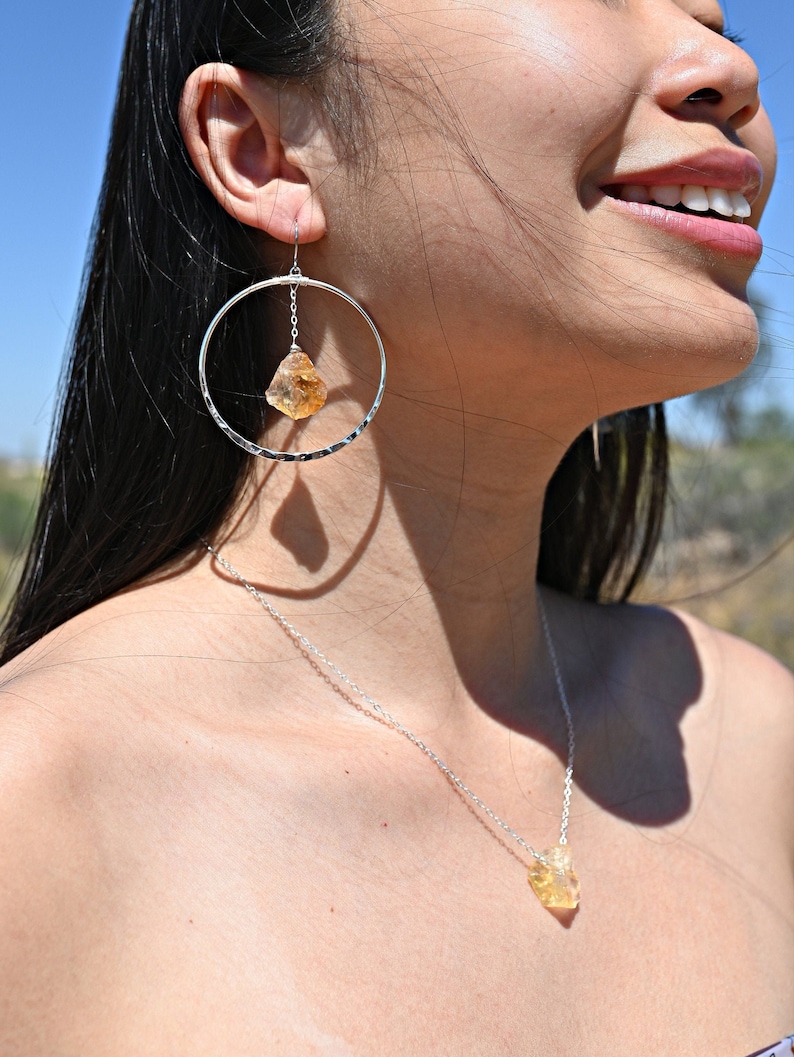 May include: A silver necklace and earrings set featuring a raw, yellow citrine stone pendant and matching earrings. The necklace is a delicate silver chain with a simple design. The earrings are hoop-style with a dangling citrine stone.