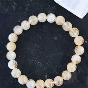 May include: A bracelet made of light yellow citrine beads. The beads are round and have a translucent appearance. The bracelet is on a dark background.