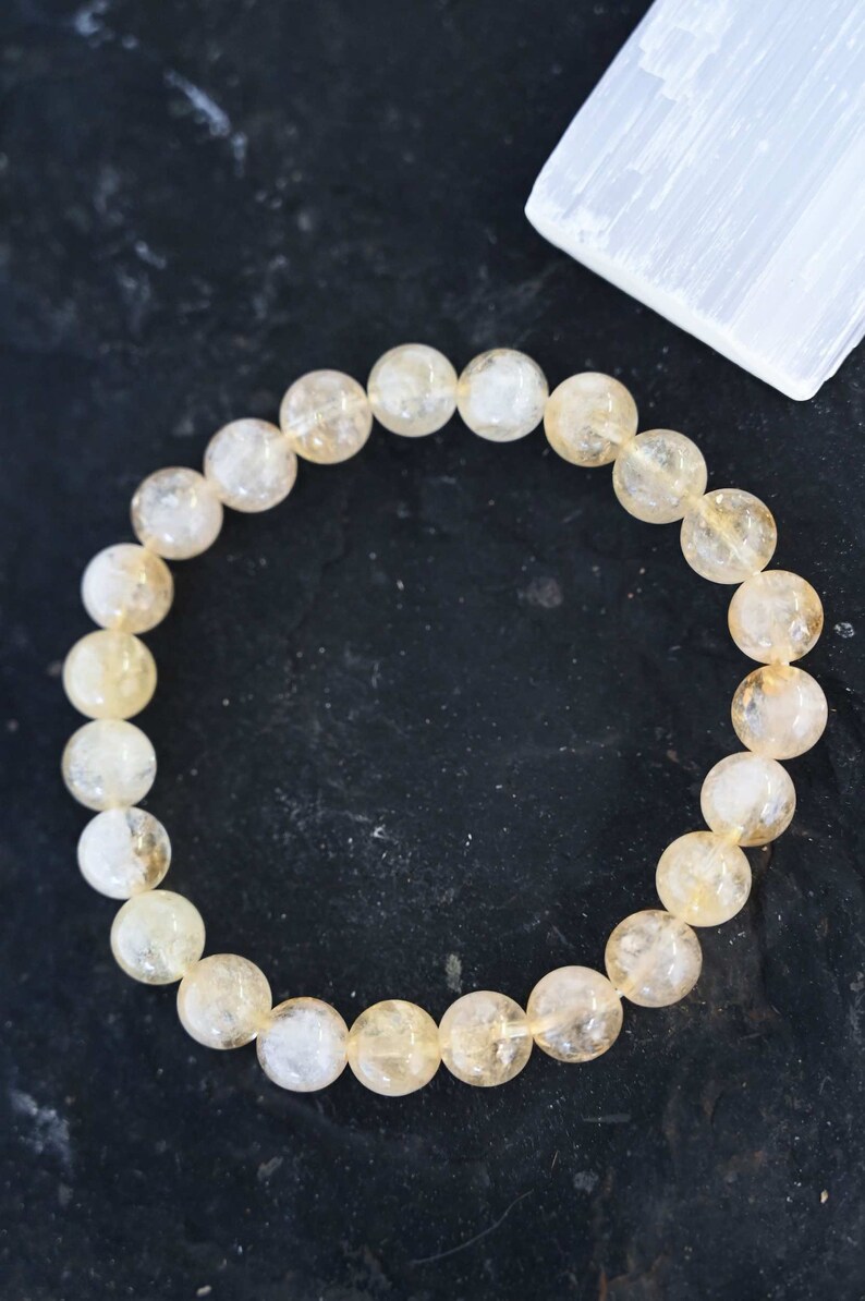 May include: A bracelet made of light yellow citrine beads. The beads are round and have a translucent appearance. The bracelet is on a dark background.